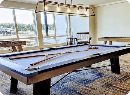 A modern game room with a pool table, two cue sticks, and a racked set of billiard balls, with large windows and natural light.